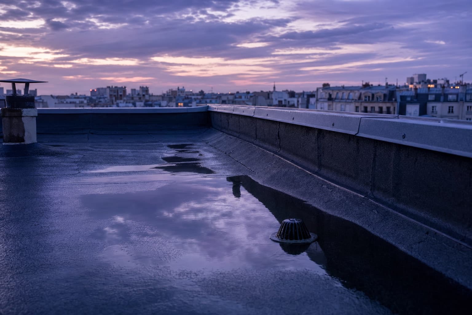 Une toiture terrasse après la pluie au crépuscule, une flaque dans un point bas, acrotère et évacuation