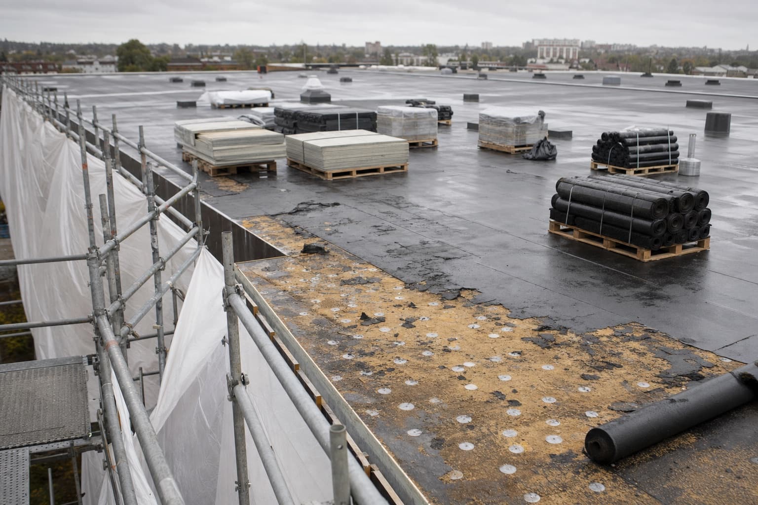 Une toiture terrasse partiellement décapée, échafaudage sur un bord et matériaux sur palettes